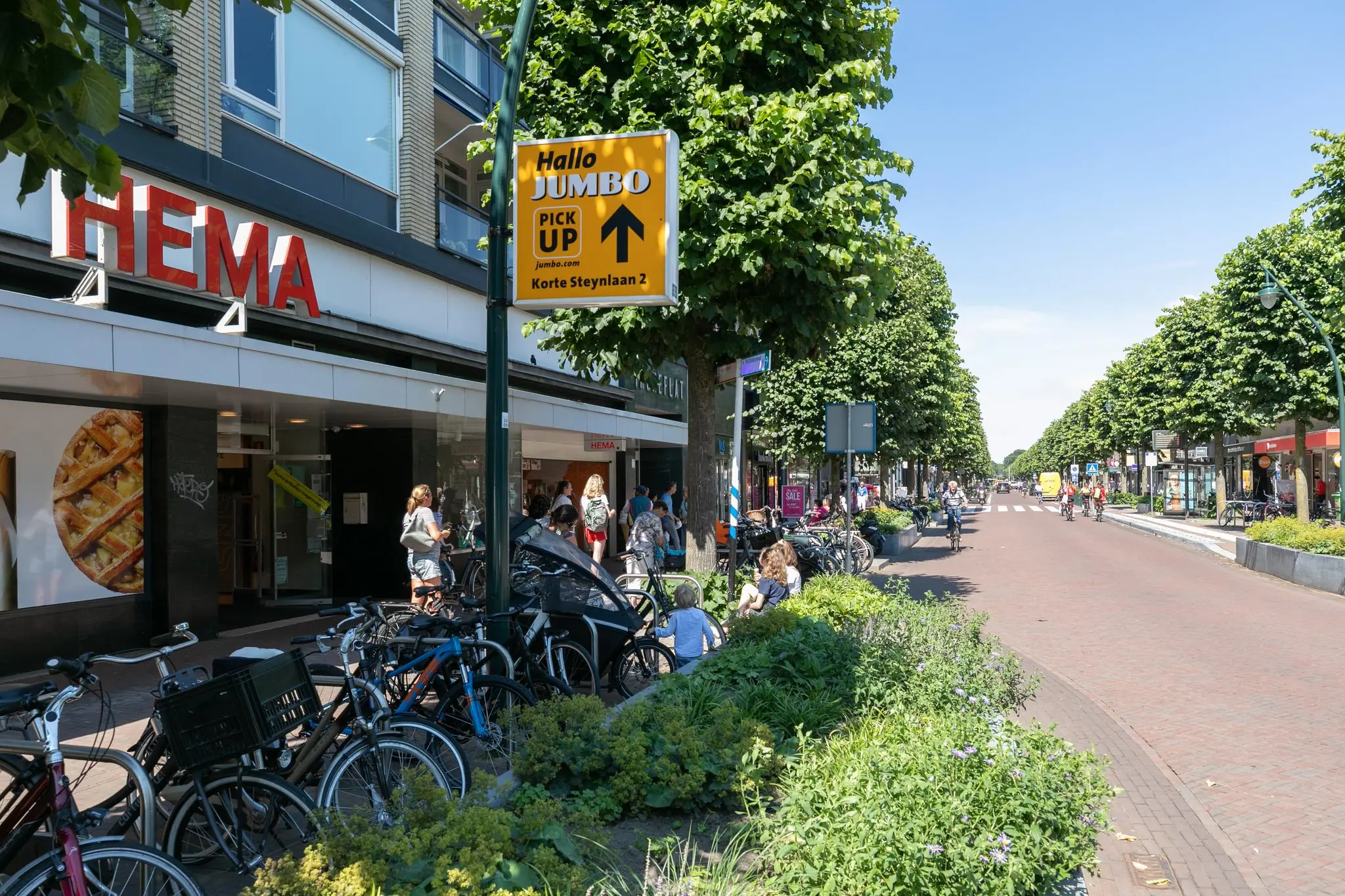 Drukke winkelstraat in De Bilt met een HEMA-filiaal, fietsenrekken en winkelend publiek op een zonnige dag.